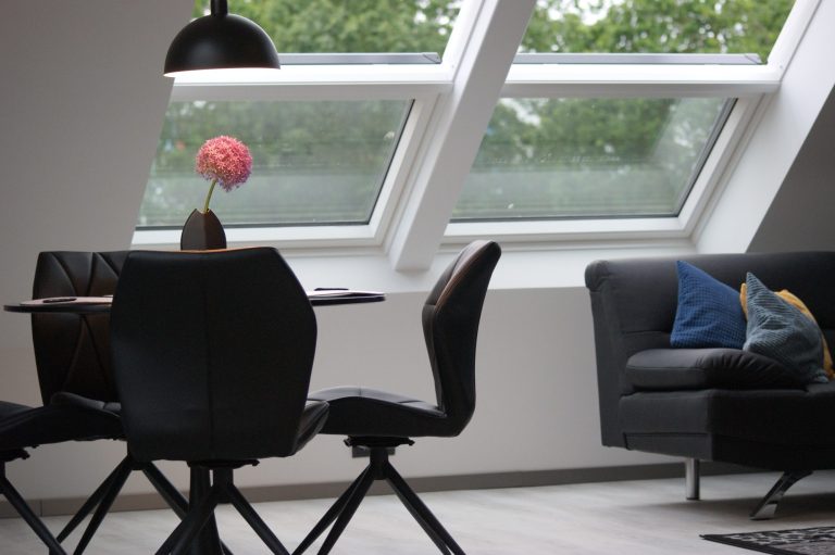 Blick auf den Tisch mit vier schwarzen Stühlen im Wohnbereich. Auf dem Tisch sind Untersetzer, helle Platzsets und eine rosafarbene Blume.