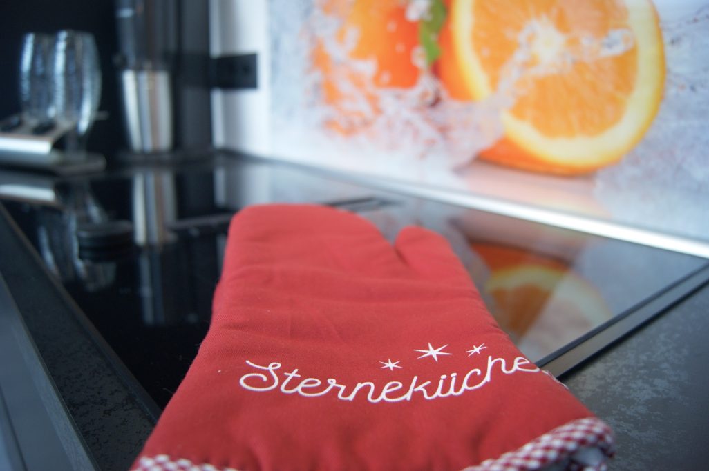 Roter Topflappen mit der Aufschrift "Sternküche" liegt auf dem Ceranfeld. Im Hintergrund ein großes Bild an der Wand mit Orangen.