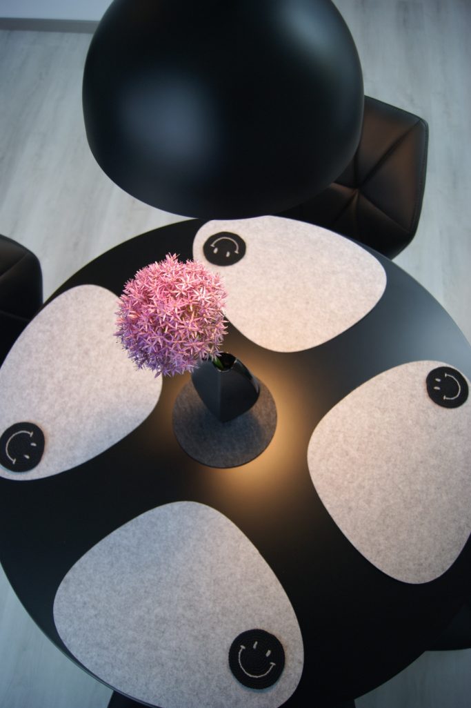 Blick von oben auf den schwarzen Tisch im Ess- und Wohnbereich mit vier hellen Platzsetzs, dunklen Untersetzern und einer rosafarbenen Blume