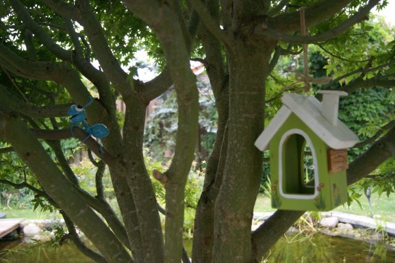 Eine blaue Ameise und ein kleines grünes Vogelhaus hängen als Deko in einem Baum am Teich.