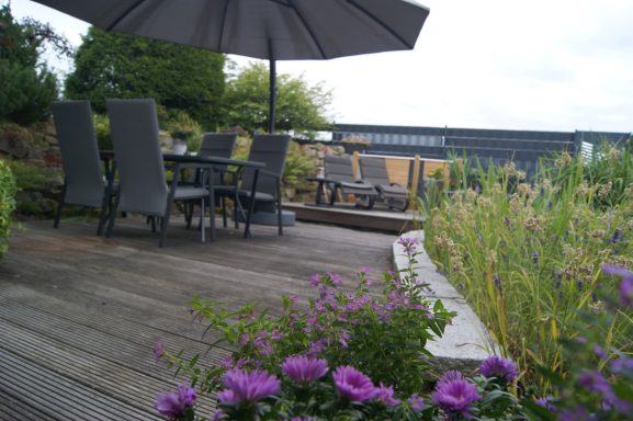 Blick auf die großzügige Terrasse der Ferienwohnung direkt am Fischteich. Vier Stühle, ein Tisch und ein Sonnenschirm sind vorne zu sehen.