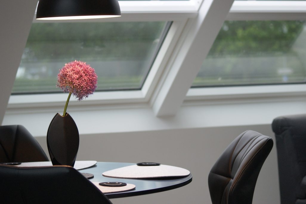 Blick auf den Tisch mit vier schwarzen Stühlen im Wohnbereich. Auf dem Tisch sind Untersetzer, helle Platzsets und eine rosafarbene Blume.